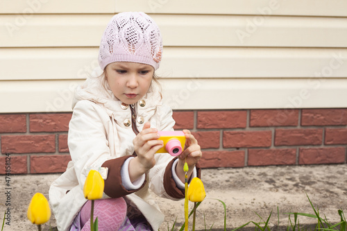 a girl in a white jacket in the spring with a children's camera sits near tulips