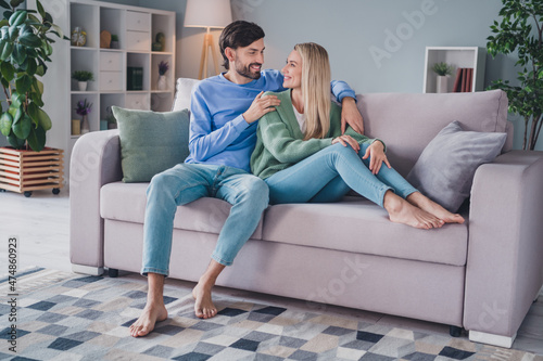 Full length photo of positive cheerful spouses sit divan man cuddle hug embrace his lady enjoy room apartment