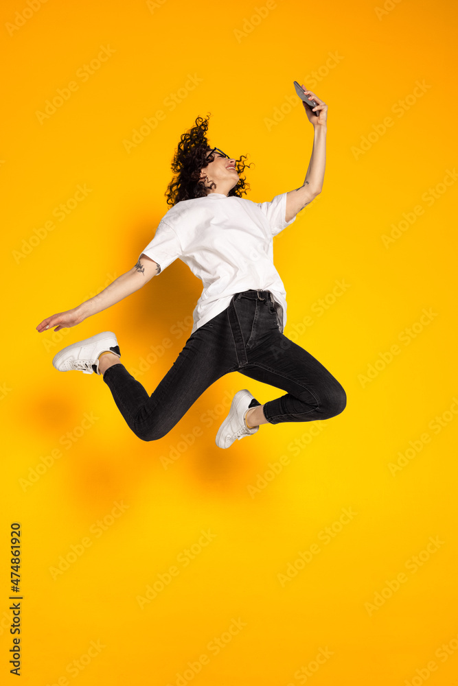Full-length portrait of young cheerful girl, student in white t-shirt and jeans jumping isolated on yellow background.