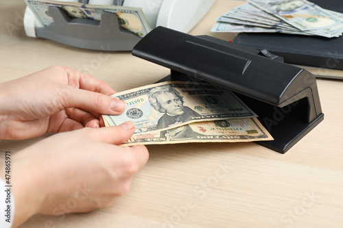Woman checking dollar banknotes with currency detector at wooden table, closeup. Money examination device