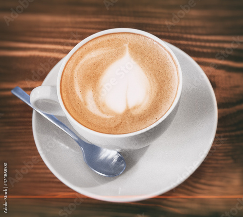 Photo of a cup with cappuccino on a white saucer with a spoon on a wooden table in a coffee shop