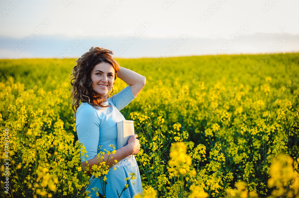 Portrait of a happy female student holding books and looking at camera outdoors. Young, beautiful girl holding a book, on a background of yellow flowers in a field Portrait of a happy female student holding books and looking at camera outdoors. Young, beautiful girl holding a book, on a background of yellow flowers in a field