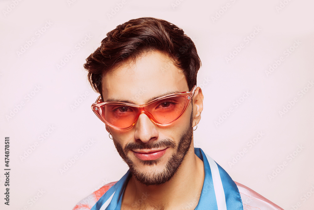 Face portrait of young smiling happy satisfied gay man 20s wear blue and pink shirt and pink stylish glasses looking at camera isolated on pastel beige background studio. Homosexual lgbtiq concept. Face portrait of young smiling happy satisfied gay man 20s wear blue and pink shirt and pink stylish glasses looking at camera isolated on pastel beige background studio. Homosexual lgbtiq concept.