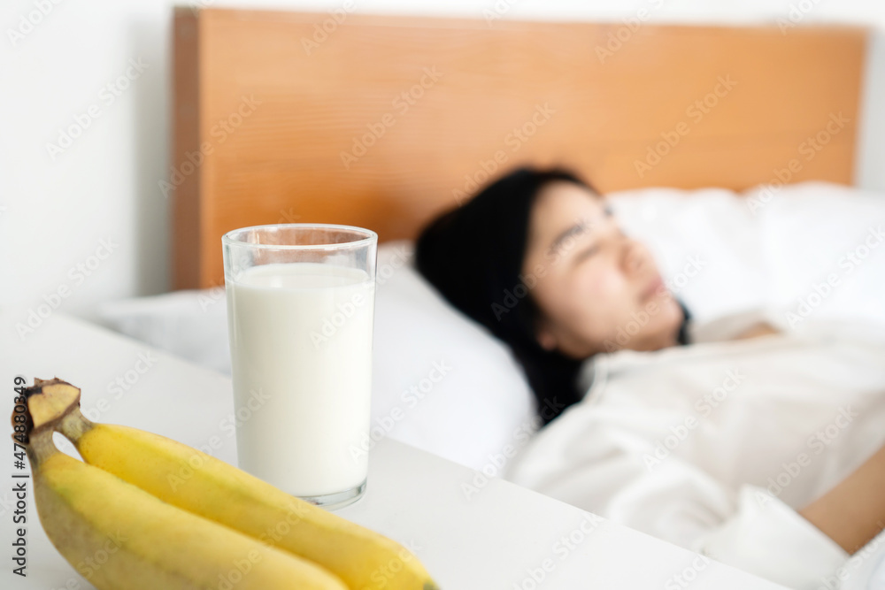 a glass of milk and bananas on the table with blur background of woman sleeping in bed, eating healthy before sleep concept
