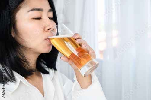 alcoholic Asian woman drinking beer hand holding a glass of beers standing next to the windows