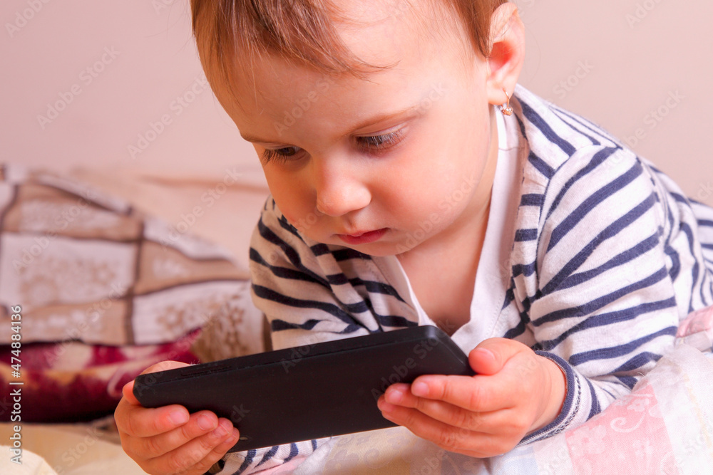 Little cute child girl using mobile phone in the bedroom before bedtime. The negative impact of gadgets on the mental state, sleep and health of the child. Horizontal image. Little cute child girl using mobile phone in the bedroom before bedtime. The negative impact of gadgets on the mental state, sleep and health of the child. Horizontal image.