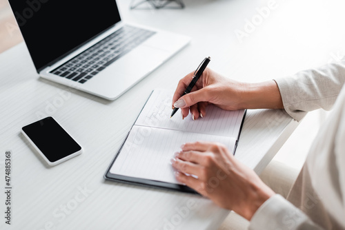partial view of businesswoman writing in notebook near gadgets