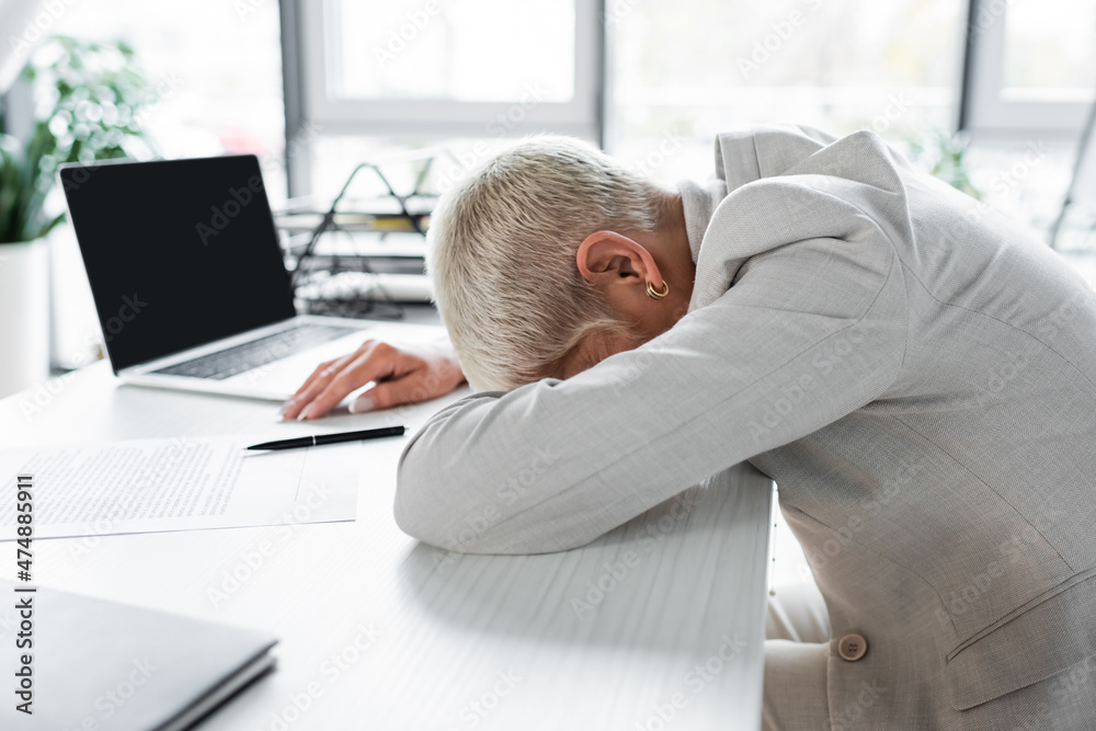tired senior businesswoman with grey hair lying on desk near laptop