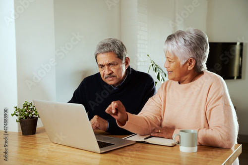 Multi-cultural elderly couple sitting at modern kitchen counter with laptop. Wife explaining to husband.