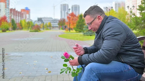 A man with a rose in his hand sits on a bench all alone in the rain and tries to light a lighter