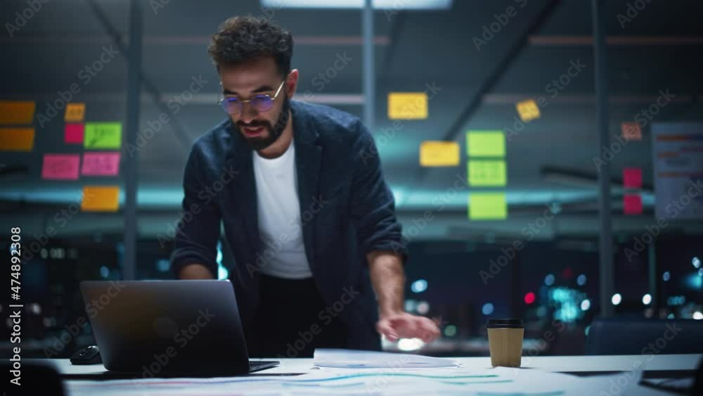 Successful Handsome Creative Director Working on Laptop Computer in Big City Office Late in the Evening. Business Analyst Preparing for a Meeting Discussion About Project Management Report.