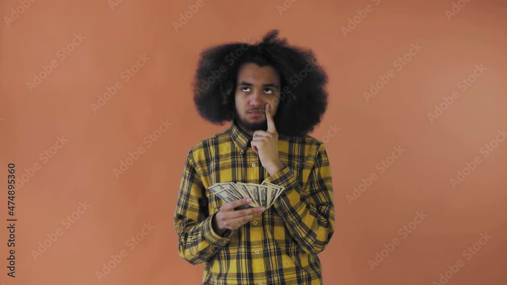 A happy African-American holding money in his hands and thinking about what to spend it on, stands isolated on an orange background A happy African-American holding money in his hands and thinking about what to spend it on, stands isolated on an orange background