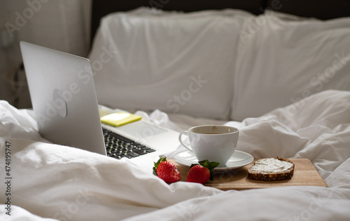 Morning coffee in bed. Cup of delicious, aromatic coffee, sandwich and two ripe strawberries on wooden kitchen board lying near laptop on white bed. Beakfast at home-office in time quarantine.