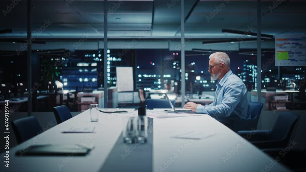 Successful Senior Businessman Working on Laptop Computer in Big City Office Late in the Evening. Investment Analyst Checking Financial Graphs from Project Management Reports. Zoom Out Shot.