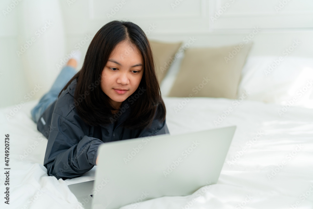 Portrait, a cute Asian teenage girl. wearing gray shirt lying in bed with a notebook computer in the bedroom to study online with a happy face Portrait, a cute Asian teenage girl. wearing gray shirt lying in bed with a notebook computer in the bedroom to study online with a happy face