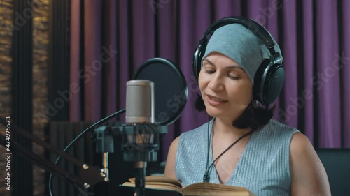 A woman in a recording studio reads aloud on a microphone an excerpt from a book. Studio recording of an audiobook. Microphone and headphones for working on audio material.