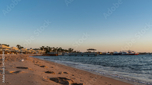 A beach on the Red Sea coast. Footprints in the sand. Umbrellas from the sun and  loungers are in a row. Above the blue water there is a path, a canopy. Boats are visible in the distance . Egypt