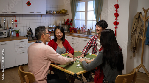 smiling asian family of four having fun with mahjong game and chatting lively in the evening at home during chinese lunar new year holiday. chinese text at background translation: luck