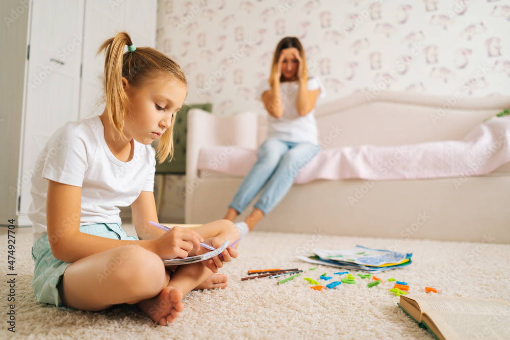 Side view of primary girl child writing homework in copybook sitting on floor with laptop for distance education online at home, on blurred background of stressed young mother sitting on sofa. Side view of primary girl child writing homework in copybook sitting on floor with laptop for distance education online at home, on blurred background of stressed young mother sitting on sofa.