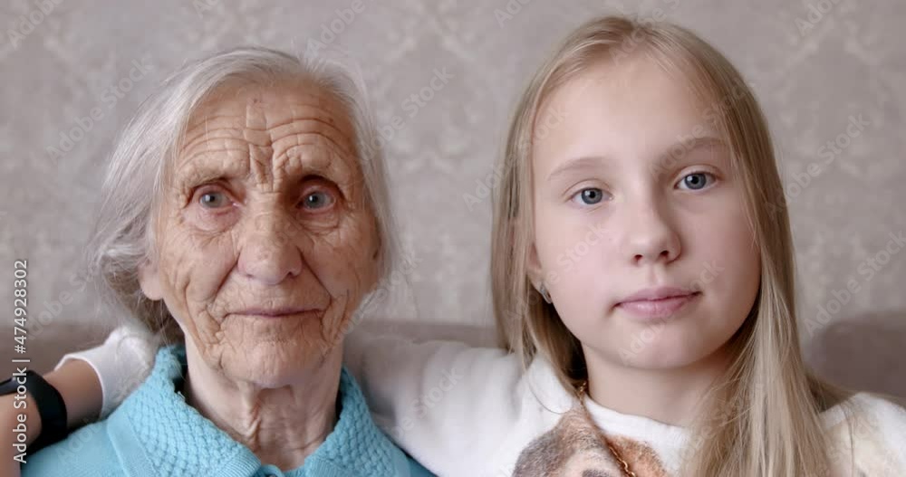 Portrait of an old woman and a teenager girl. The face of a grandmother with deep wrinkles and a young girl are sitting together on the couch at home. 