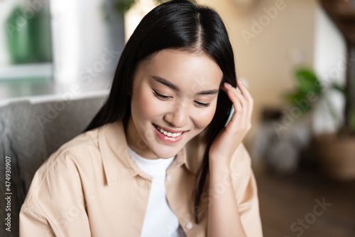 Cute asian girl student smiling, looking down and tucking hair behind ear, sitting at home indoors in casual clothes photo