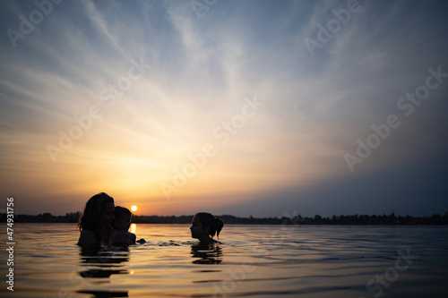 Mom with the baby she holds in her arms playing with her daughter and splashing in the lake against background of sunset © YouraPechkin