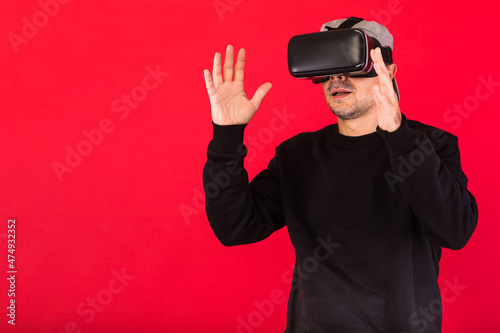 Young man with short beard, wearing black sweatshirt, cap and virtual reality glasses amazed, touching something virtually, on red background. Technology, VR, computing and hobbies concept. © Davidbenito