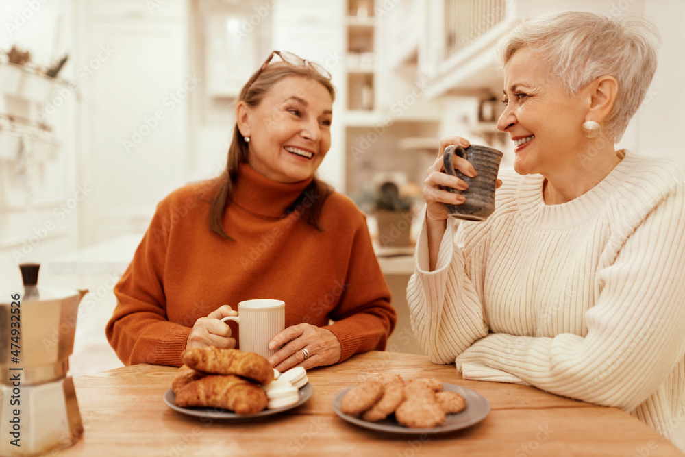 © shurkin_son - People, lifestyle and leisure concept. Portrait of stylish senior women in cozy sweaters having breakfast together at cafe, sitting at wooden table, sharing news, drinking coffee with pastries
