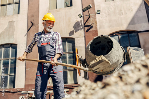 A senior construction worker is putting cement into a cement blender while standing at the construction site. The builder is mixing cement and preparing for the build. A worker with a cement blender.