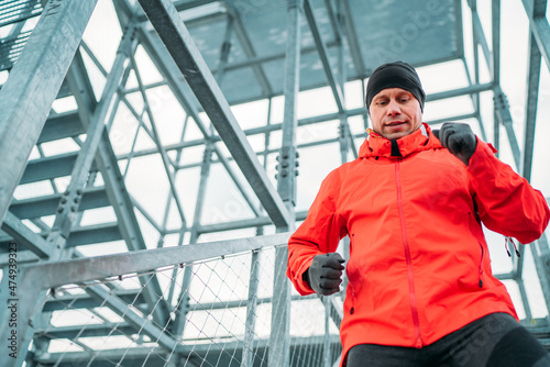 Waist portrait of smiling runner man dressed bright red softshell sporty clothes and gloves running down by huge steel industrial stairs in cold winter day. People healthy lifestyle concept photo. photo