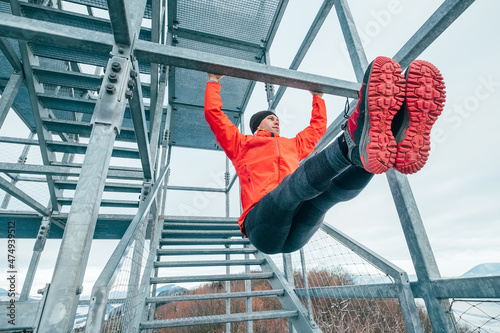 A fit man dressed in bright red sporty clothes and running shoes hanging horizontal bar training abs by raising legs. Fitness workout on the steel industrial construction.