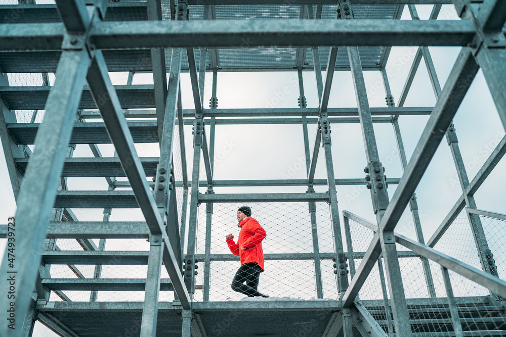 Alone runner man dressed bright red softshell sporty clothes running up by huge steel industrial stairs in cold winter day. People healthy lifestyle concept photo.
