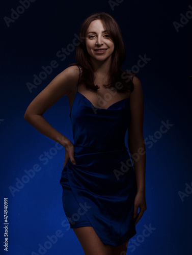 Young and cute brunette woman in blu dress posing on dark blue background free space for text