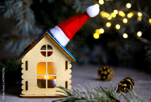 selective focus on a small wooden house, a candle will burn inside, a santa cap on the roof, against the background of blurred specks of garland, the coneption of Christmas and New Year photo