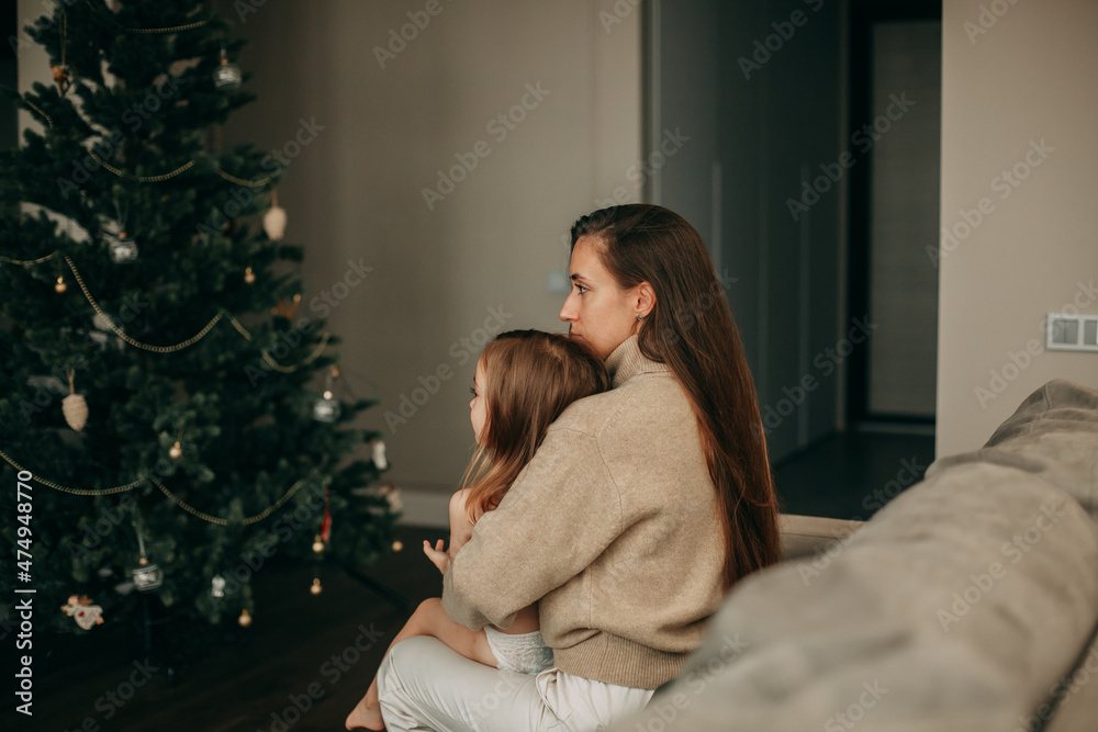 Mom sits on the couch at home and holds her daughter in her arms. Against the background of a Christmas tree