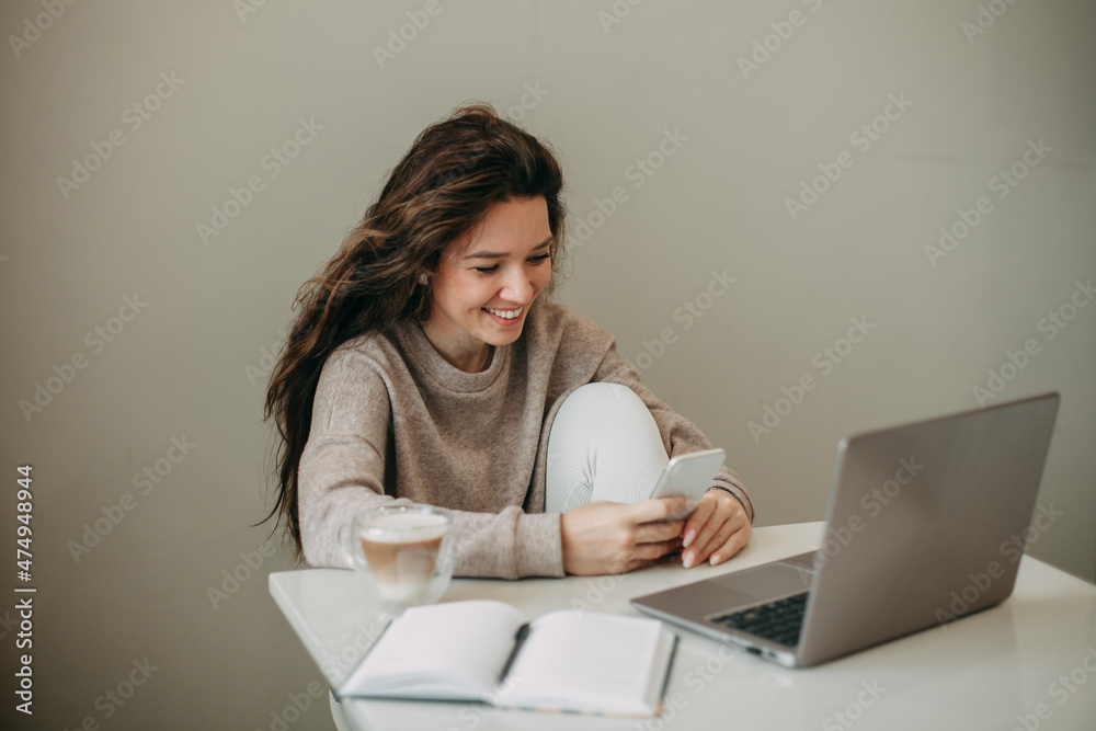 young brunette woman 30 years old with long hair sits at home and uses a smartphone. On a white table is a mug of coffee, a notebook with a pen, a laptop. young brunette woman 30 years old with long hair sits at home and uses a smartphone. On a white table is a mug of coffee, a notebook with a pen, a laptop.