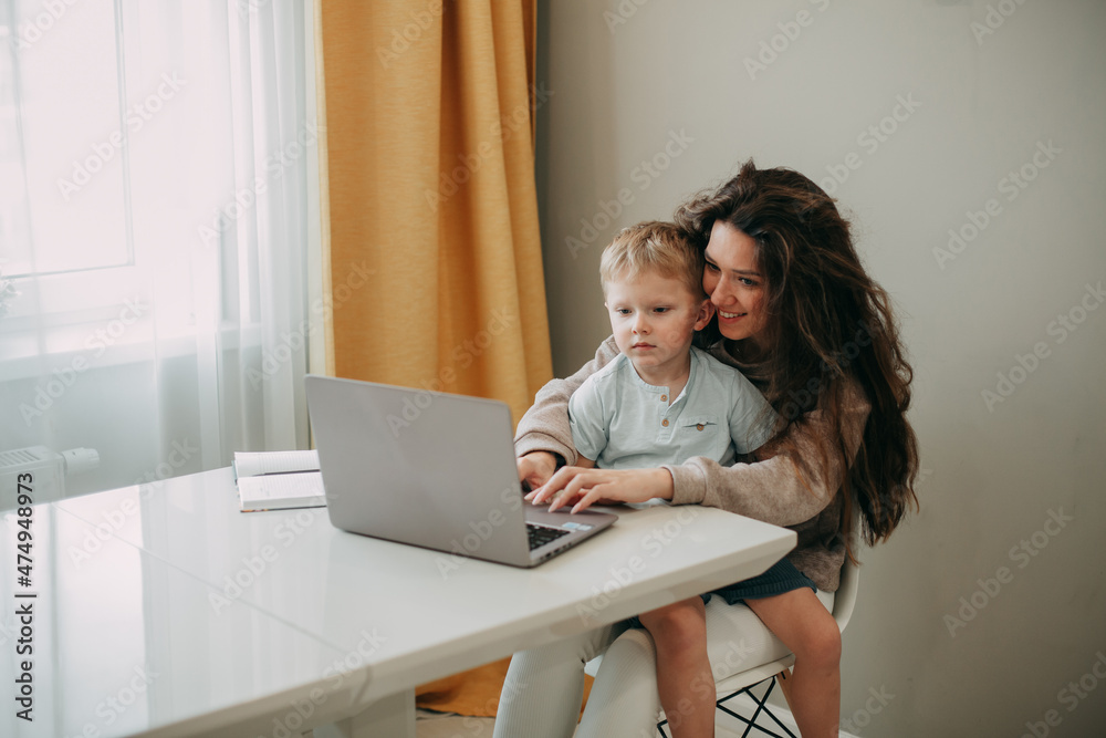 Smiling young 30 years old brunette woman with long hair sits on a chair at home. A son is sitting in her arms. Look at the laptop screen. Smiling young 30 years old brunette woman with long hair sits on a chair at home. A son is sitting in her arms. Look at the laptop screen.