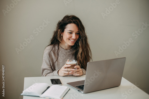 Smiling young 30 years old brunette woman with long hair uses laptop at home. He holds a mug of coffee in his hands, looks at the laptop screen
