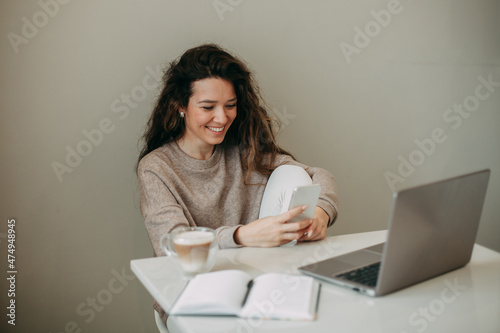 young brunette woman 30 years old with long hair sits at home and uses a smartphone. On a white table is a mug of coffee, a notebook with a pen, a laptop.