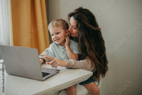 Smiling young 30 years old brunette woman with long hair sits on a chair at home. A son is sitting in her arms. Look at the laptop screen.