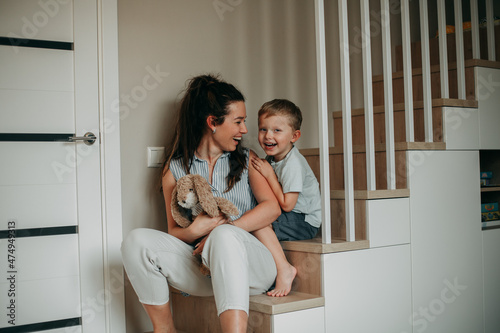 A young girl of 30 years old brunette with long hair in a white shirt with blue stripes and white jeans fooling around at home with her blond son in a blue shirt and blue shorts