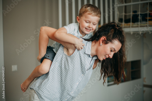A smiling brunette mom 30 years old with long hair in a white shirt with blue stripes and white jeans at home rolls her son on her back. Blond boy in a blue t-shirt and blue shorts