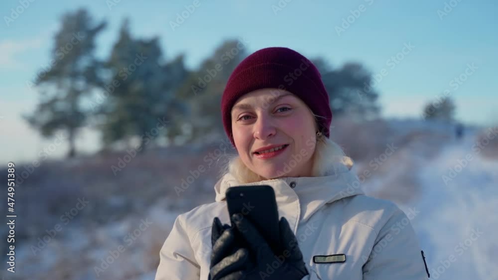 Portrait female holding smartphone looking at camera at winter woods outdoors. Attractive woman standing at forest outside and smile. Girl dressed in warm clothes. Slow motion Portrait female holding smartphone looking at camera at winter woods outdoors. Attractive woman standing at forest outside and smile. Girl dressed in warm clothes. Slow motion