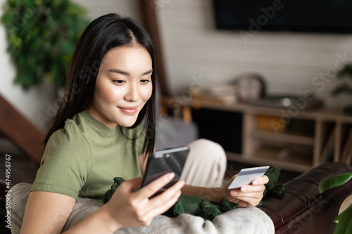 Smiling korean girl using mobile phone and credit card while sitting at home, paying for purchase in internet. Buying something from her house photo