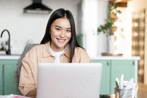 Young asian girl freelancer working remotely from home. Woman with laptop studying indoors from her kitchen, smiling at camera
