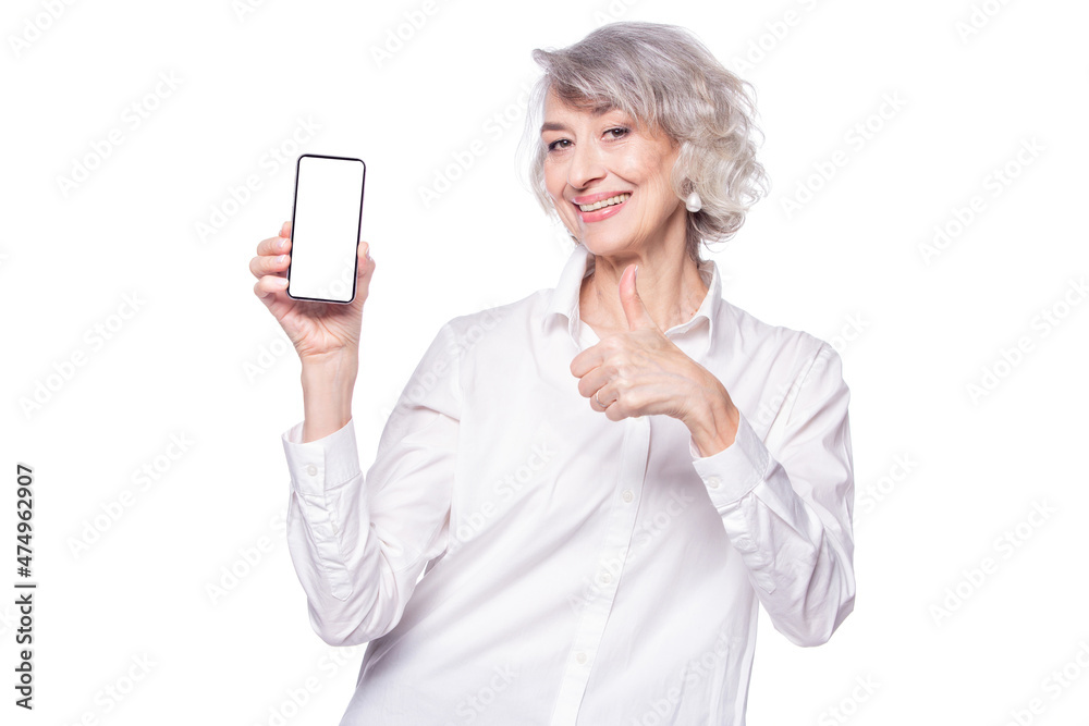 A beautiful happy modern senior woman looks at the camera with a wide smile showing a smartphone in her raised hand and showing her thumb up expressing approval, isolated on white background A beautiful happy modern senior woman looks at the camera with a wide smile showing a smartphone in her raised hand and showing her thumb up expressing approval, isolated on white background