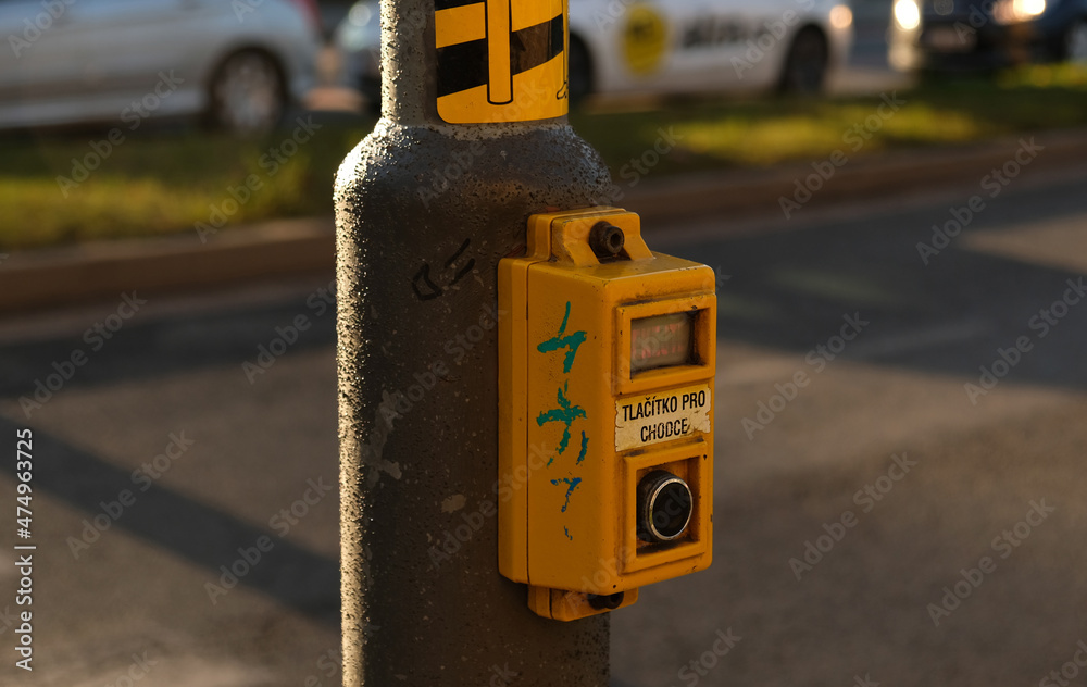 Crosswalk button for pedestrian with light warning. Defocused background with Cars drive along road and bokeh from headlights. The inscription in Czech -Tlacitko pro chodce- Button for pedestrians. 