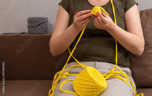 Woman is wrapped in yarn, holds hook in her hands and knits product from yellow knitted yarn. Place for your text. Selective focus. Picture for articles about knitting, people's hobbies.