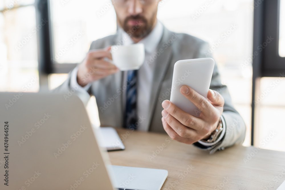 Cropped view of smartphone in hand of blurred businessman holding cup near laptop in office. Cropped view of smartphone in hand of blurred businessman holding cup near laptop in office.