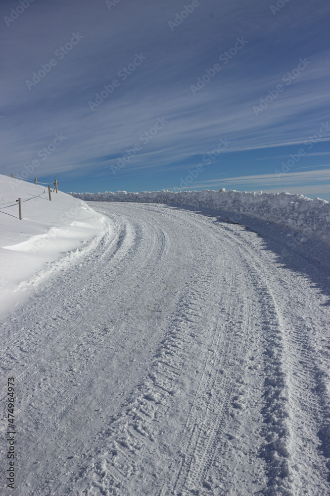 Beautiful winter mountain road covered with snow against the sky with long white clouds. Snowdrifts left on the roadside. Treviso Prealps, Italy. Vertical image.